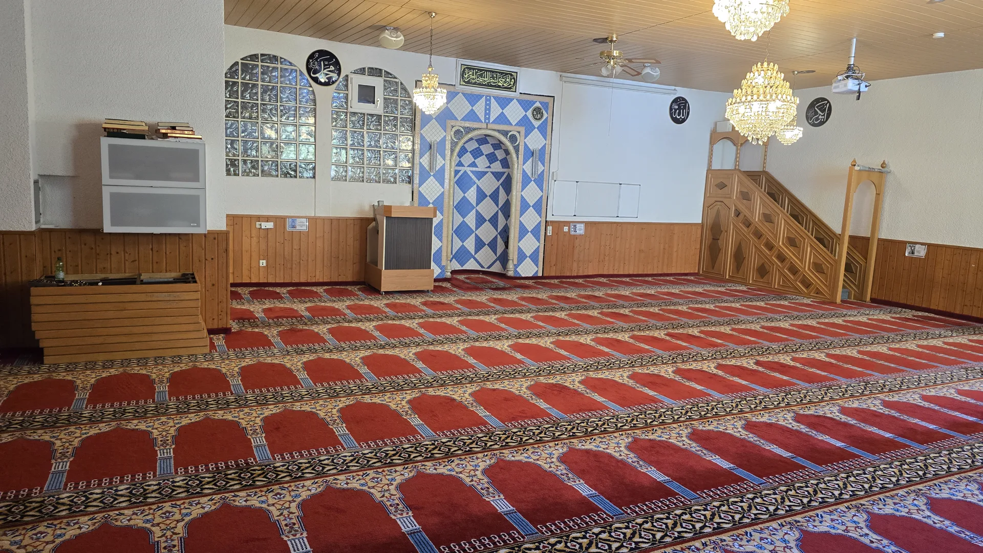 Der Gebetsraum der Moschee: roter Gebetsteppich in Reihen, die blau geflieste Gebetsnische, der hölzerne Minbar und Kronleuchter